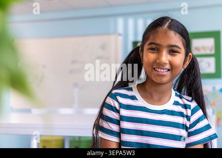 Indian child smiling, facing camera at school by whiteboard in striped tee, copy space Stock Photo