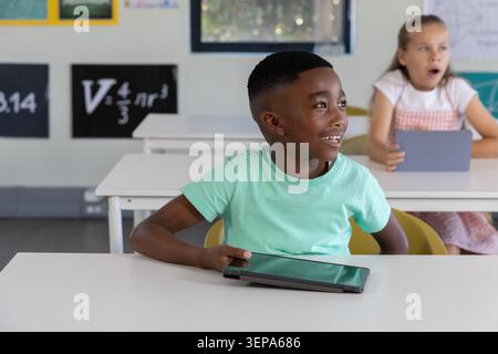 Two classmates sitting in classroom, boy smiling, girl in pink-gingham holding tablet, math posters Stock Photo