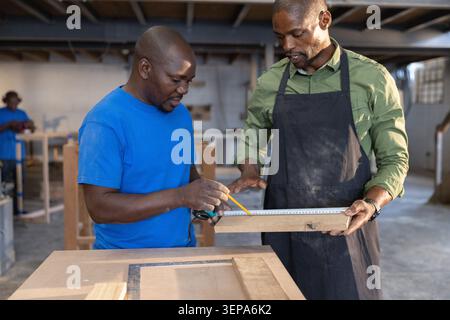 African American coworkers in aprons measuring  and  marking plank using tape measure  and  pencil i Stock Photo