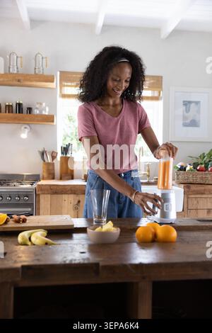 Smiling African American woman blending orange smoothie on kitchen island with blender and fruits Stock Photo