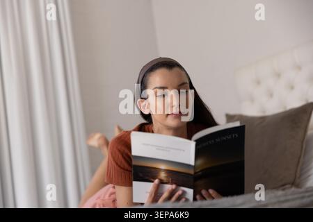 Female lying on stomach reading book on bed, wearing rust top and brown headband Stock Photo