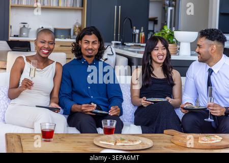Diverse friends sitting in living room on light sofa sharing pizza and sipping drinks Stock Photo