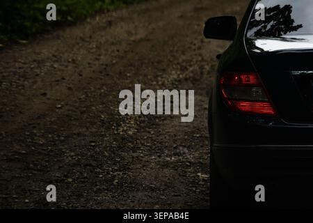 Right taillight of a dark sedan glows beside a winding gravel path through dense woods with wet pebbles scattered on the surface creating a moody trav Stock Photo