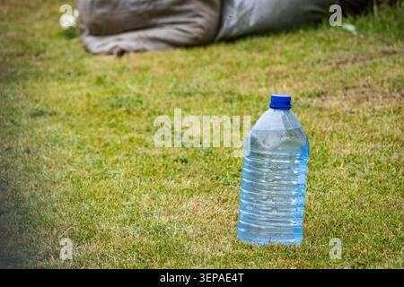 Large clear plastic water jug with blue cap standing on trimmed lawn in garden. Sunlight catches ripples through transparent surface. Simple outdoor h Stock Photo