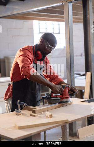 Red power sander smoothing long wooden plank on worktable inside workshop, with wood offcuts Stock Photo