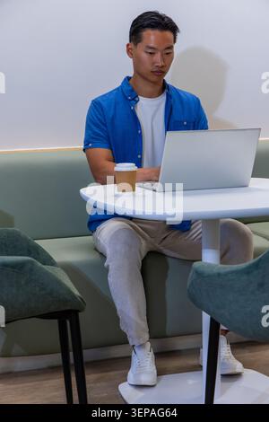 Asian man working on silver laptop at cafe on light green bench, with paper coffee cup Stock Photo
