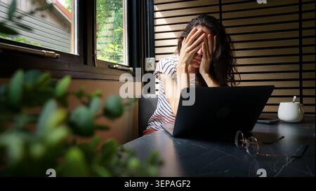 Woman feeling stressed and exhausted covering her face while working remotely on a laptop Stock Photo
