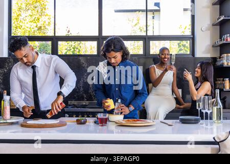 Diverse friends making pizza on kitchen island, man in tie sprinkling sauce, woman using smartphone Stock Photo