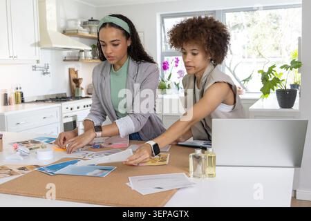 African American women in blazer and vest arranging corkboard prints on kitchen island using laptop. Collaboration, design, brainstorming, moodboard, Stock Photo