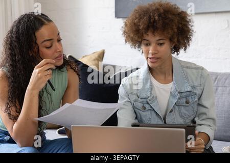 African American women friends sitting on gray sofa reviewing papers and using laptop with tablet Stock Photo