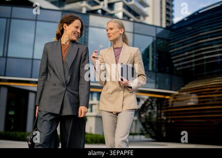 Two women walk together outside a modern building in the city. They discuss something while holding documents and smile at each other. The scene is bu Stock Photo
