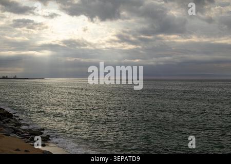 Wide coastal view with sunlight streaming through dense clouds onto a calm sea. Rocky shoreline and gentle waves create a peaceful maritime scene with Stock Photo