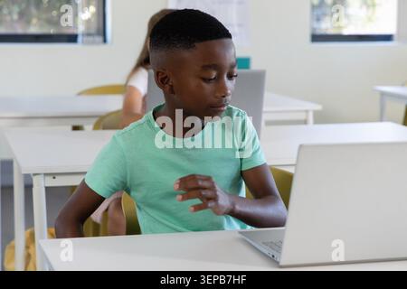 Diverse child classmates sitting at white desks in classroom using silver laptop and mint shirt Stock Photo