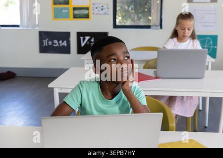 Diverse children sitting at desks on laptops, African American boy resting cheek wearing green tee Stock Photo