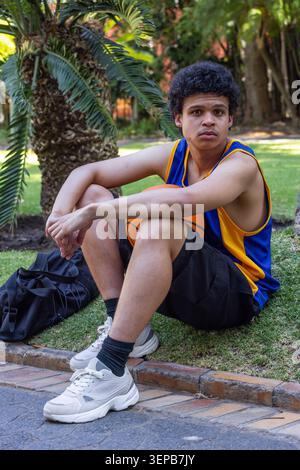 Adult African man sitting on grass in park holding orange basketball in blue jersey, black backpack Stock Photo