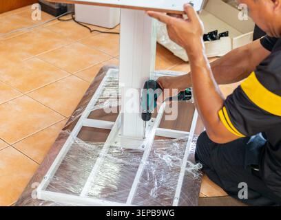 Close-up of a worker assembling office furniture, fixing a white metal table leg structure with a power tool during home installation service. Stock Photo