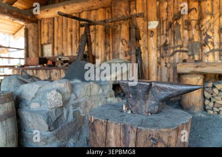 Rustic forge interior with heavy anvil on wooden stump, stone hearth, hanging tools, rough timber walls, and warm natural light inside an old workshop Stock Photo