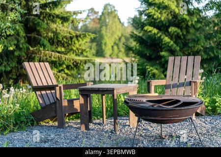 Rustic seating area with two wooden chairs, a small table, and a metal fire pit on gravel near a calm lake, surrounded by green grass, evergreen trees Stock Photo