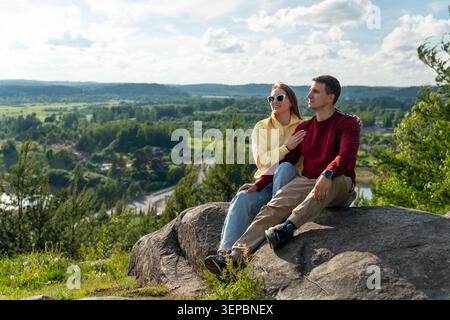 A young man and woman sit together on a large rock above a scenic green valley, enjoying the open landscape, bright daylight, and calm outdoor setting Stock Photo