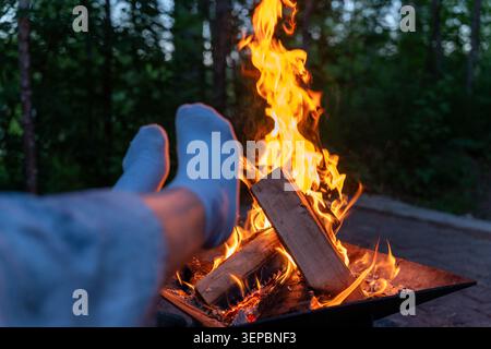 A close outdoor scene shows glowing campfire burning in a metal fire pit with a person resting their feet nearby background features wooded area Stock Photo