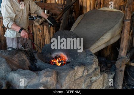 A worker in protective gloves tends a hot coal forge inside a rustic wooden workshop, with tools and an anvil surface lit by warm firelight Stock Photo