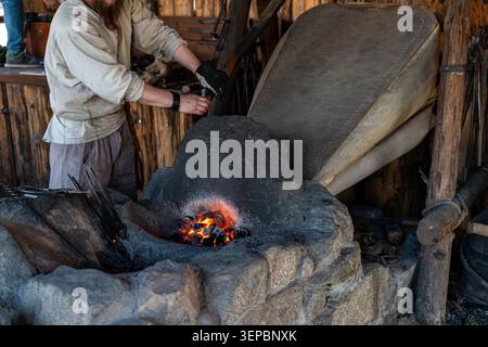 Person in protective gloves tends hot metal at a rustic forge with glowing embers heavy tools, and weathered workshop interior made of wood and stone Stock Photo