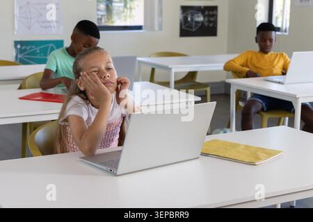 Diverse children sitting at spaced white desks in classroom using silver laptops and yellow chairs Stock Photo