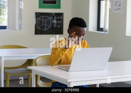 African American male child wearing mustard top sitting at desk using laptop pointing in school Stock Photo