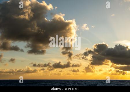 Dramatic golden clouds illuminated by the setting sun over a calm ocean, with sun rays visible through breaks in the clouds under a clear sky. Stock Photo