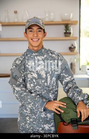 Asian man in camouflage uniform standing and smiling in kitchen, resting hands on green duffel bag Stock Photo