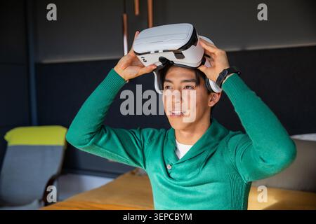 Adult Asian man sitting at wooden table in lounge wearing green sweater holding white VR headset Stock Photo