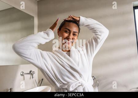 Asian adult woman adjusting hair, smiling in plush white bathrobe in bathroom with chrome faucet Stock Photo