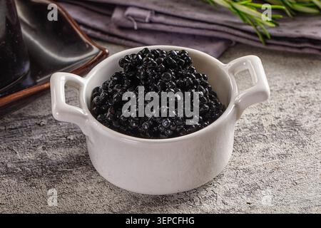 Close up of black caviar in white ceramic bowl with handles on textured gray surface with rosemary sprig and dark napkin in background Stock Photo