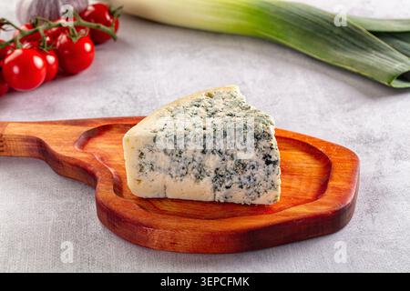 Wedge of blue cheese on rustic wooden serving board, accompanied by cherry tomatoes on vine and fresh leek on light grey stone background, gourmet foo Stock Photo