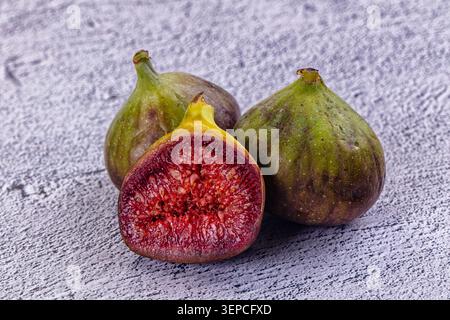 Three fresh green figs on gray textured surface. One fig is cut in half revealing vibrant red interior with seeds. Natural lighting highlights fruit d Stock Photo