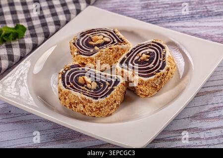 Three slices of sweet pastry rolls with dark filling, sesame seeds, and walnuts on white plate with checkered cloth Stock Photo