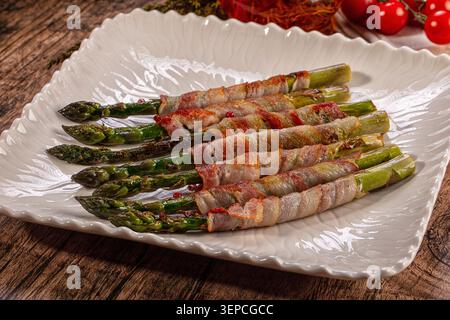 Closeup of grilled green asparagus spears wrapped in crispy bacon served on a white ceramic platter with cherry tomatoes in background. Rustic wooden Stock Photo