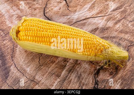 Bright yellow corn on the cob with husk partially peeled back, resting on textured wood grain surface. Close up food photography highlighting natural Stock Photo