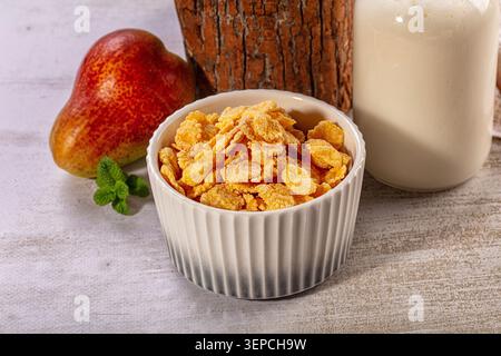 Golden cornflakes in fluted white ceramic bowl, fresh pear, mint sprig, glass bottle of milk, rustic wood background, healthy breakfast concept, warm Stock Photo