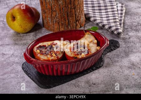 Delicious baked pear halves in red ceramic dish, topped with golden crumble pine nuts. Warm, inviting dessert on rustic grey surface with fresh pear c Stock Photo