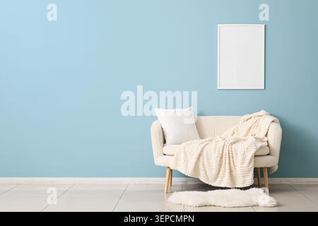 Cozy living room interior with cream sofa, soft blanket, white pillow, and blank picture frame on blue wall Stock Photo