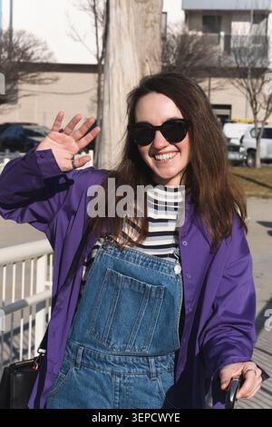 Young woman smiling and waving her hand at the camera outdoors Stock Photo