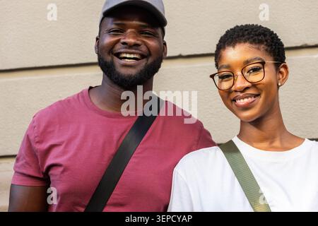 African American couple standing smiling against beige panel wall, crossbody straps and eyeglasses Stock Photo