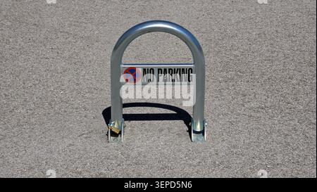 A silver metallic u shaped folding parking barrier featuring a bold no parking sign and a small padlock stands secured upright on a gray textured Stock Photo