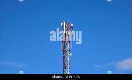 A tall red and white metal lattice telecommunication tower equipped with multiple white panel antennas and a small satellite dish stands under a vibra Stock Photo