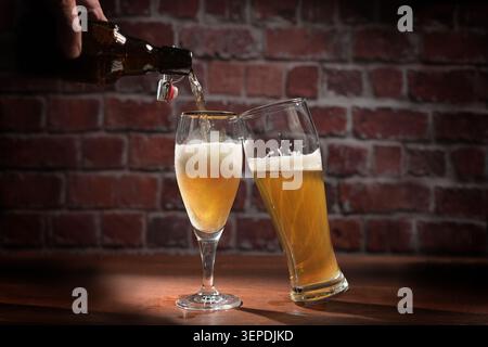 Beer is poured from a bottle into one of two glasses, refreshing alcoholic drink on a wooden table against a dark, rustic brick wall in a bar, copy sp Stock Photo