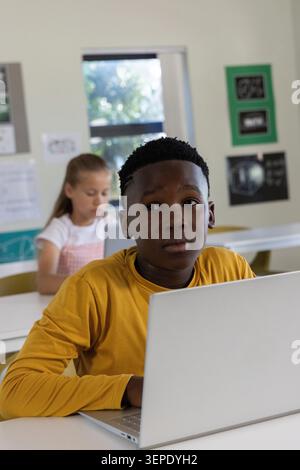 Diverse classmates sitting at classroom desks, boy mustard shirt using silver laptop, girl writing Stock Photo