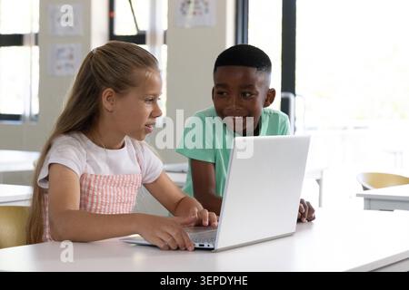 Diverse children sitting at white table using silver laptop and notebook in bright classroom Stock Photo