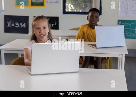 Diverse school kids sitting in classroom using laptops near math posters, girl in white shirt. Students, computers, education, geometry, diagrams, day Stock Photo