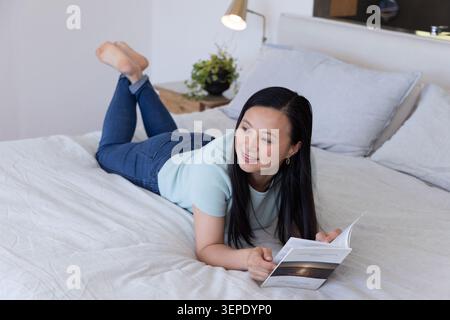 Mature Asian woman lying on stomach on bed reading paperback, wearing blue top and jeans Stock Photo